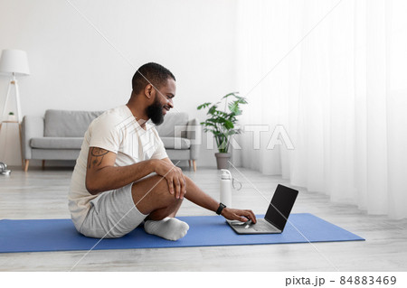 Happy young black man on floor on mat in living room interior with bottle of water, looks at laptop Happy young black man on floor on mat in living room interior with bottle of water, looks at laptop 84883469