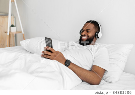 Cheerful young african american man with beard in headphones, typing on smartphone, enjoy listening music Cheerful young african american man with beard in headphones, typing on smartphone, enjoy listening music 84883506