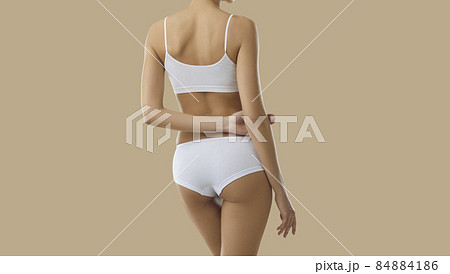 Cropped shot of young female model in underwear. Back view of beautiful Caucasian woman with fit, attractive body wearing basic soft white bra and underpants posing isolated on solid beige background 84884186