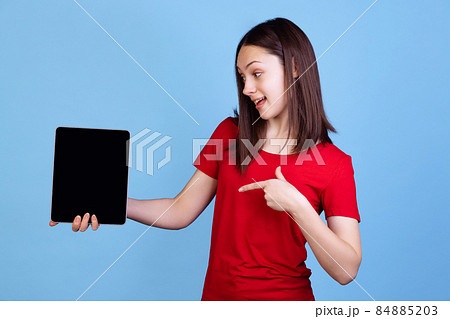 Emotional young beautiful girl in red t-shirt using digital tablet isolated on blue studio background. Concept of emotions 84885203