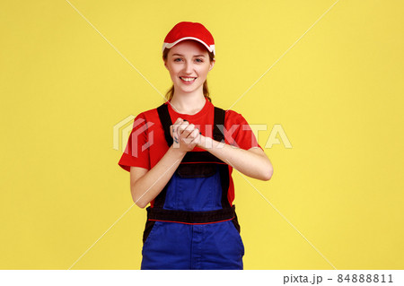 Portrait of smiling handy woman pressing hands together on chest enjoying, calming and comforting herself, wearing overalls and red cap. Indoor studio shot isolated on yellow background. Portrait of smiling handy woman pressing hands together on chest enjoying, calming and comforting herself, wearing overalls and red cap. Indoor studio shot isolated on yellow background. 84888811
