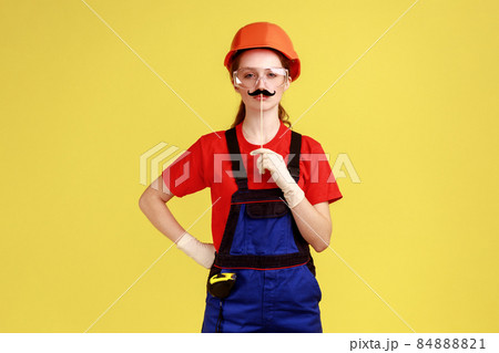 Funny confident builder woman standing with mustache on stick, looking at camera, holding hand on chin, wearing overalls and protective helmet. Indoor studio shot isolated on yellow background. 84888821