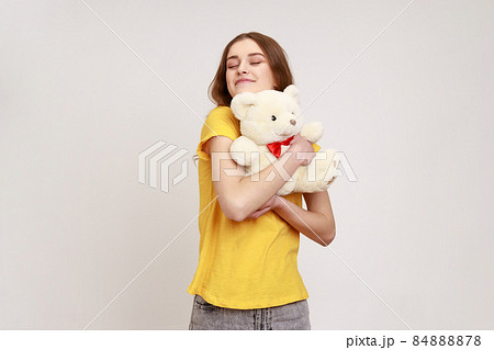 Satisfied happy teenager girl with brown hair in yellow casual T-shirt hugging cute white toy bear, closing eyes and dreaming, enjoying romantic gift. Indoor studio shot isolated on gray background. 84888878