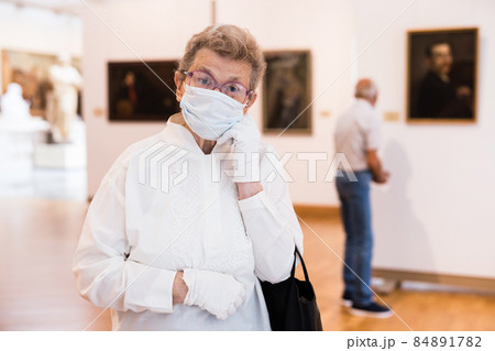 elderly European woman in mask protecting against covid examines paintings on display in hall of art museum elderly European woman in mask protecting against covid examines paintings on display in hall of art museum 84891782