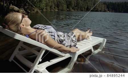 Woman lie on a sunbed in sunglasses and a boho silk shawl. Girl rest on a flood wood underwater pier. Ducks and swans are swimming nearby. 84895244