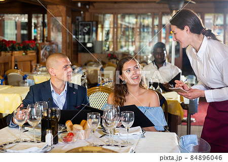 Young positive waitress taking table order from guests Young positive waitress taking table order from guests 84896046