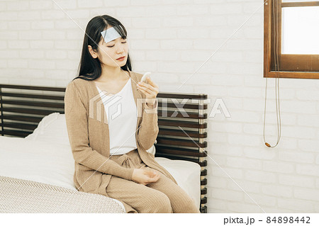 冷却シートとパジャマでベッドに座りながら体温計で体温を測る体調不良の女性 84898442