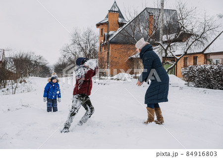 Outdoor family activities for happy winter holidays. Happy mother and two sons playing snowballs on snowy street in suburb. Happy family on winter weekend, Christmas holidays 84916803