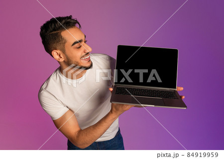 Millennial Arab man showing laptop computer with empty black screen in neon light, mockup for new website 84919959
