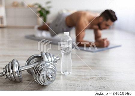 Young Arab guy standing in elbow plank pose on yoga mat at home, focus on dumbbells and bottle of water, copy space 84919960