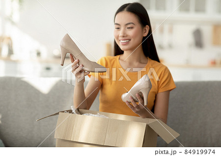 Satisfied asian woman unpacking cardboard box with new shoes, looking at high heels and smiling, sitting on sofa at home Satisfied asian woman unpacking cardboard box with new shoes, looking at high heels and smiling, sitting on sofa at home 84919974