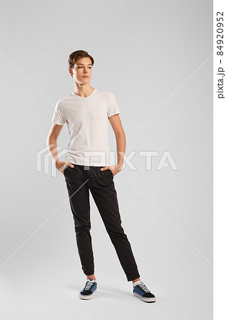 Nice man in white t-shirt, black trousers and sneakers posing over grey background Nice man in white t-shirt, black trousers and sneakers posing over grey background 84920952