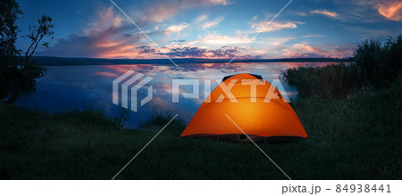Internally lit orange tent on shore of lake under dramatic sunset sky Internally lit orange tent on shore of lake under dramatic sunset sky 84938441