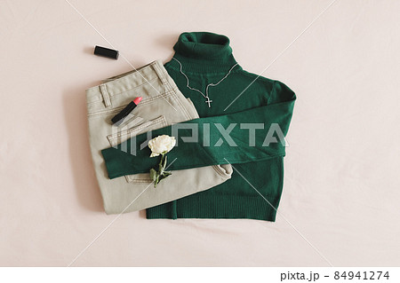 Overhead view of woman's casual outfit. Beige jeans, green knitted sweater, cosmetics and accessories lie on beige background. Trendy stylish women clothes. Flat lay, top view 84941274