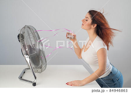 A red-haired Caucasian woman chills by the electric fan and drinks a cold drink. Climate control in the apartment 84946863