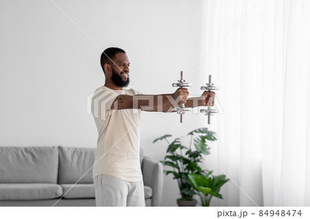 Happy young african american male with beard in white sportswear raises dumbbells in living room 84948474
