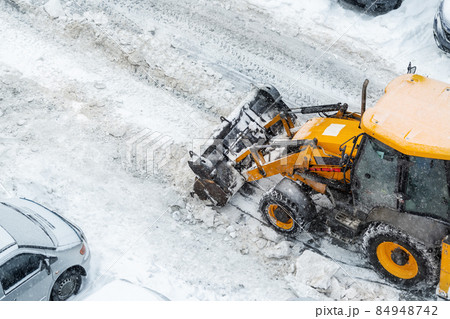 Tractor loader machine uploading dirty snow into dump truck. Cleaning city street, removing snow and ice after heavy snowfalls and blizzard. Snowplow outdoors clean pavement sidewalk road driveway 84948742