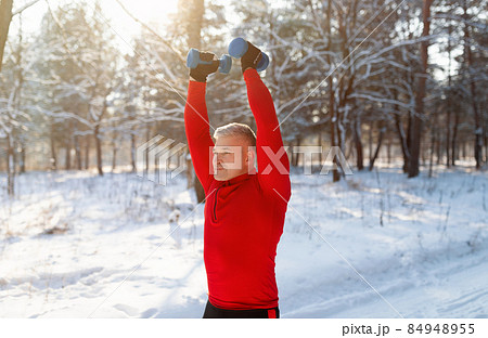 Outdoor winter sports. Athletic senior man working out with dumbbells, training arm muscles at snowy forest 84948955