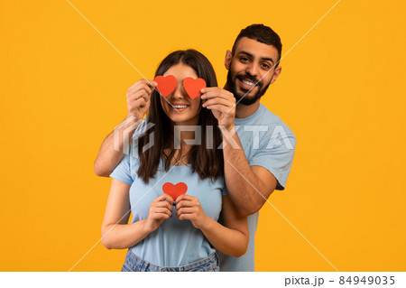 I love you. Young loving arab man covering woman eyes with surprise and giving her heart cards, yellow background 84949035