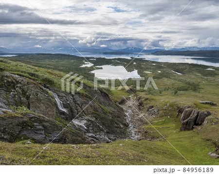 Lapland landscape at Virihaure lake with small waterfall, green mountains, birch trees and plain with big boulders. Sweden summer moody and foggy wild nature, Padjelantaleden hiking trail. 84956189