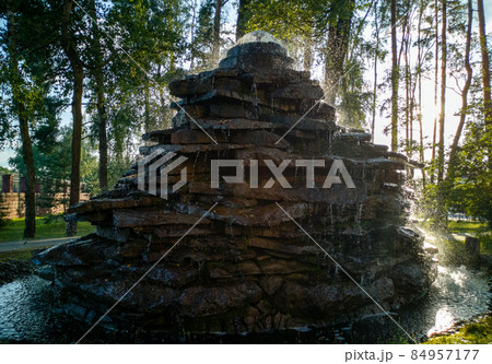 Close up on a stone made structure resembling a natural well or waterfall with a bamboo stick used for pouring water seen in the middle of a public park with many shrubs and trees in the background. Close up on a stone made structure resembling a natural well or waterfall with a bamboo stick used for pouring water seen in the middle of a public park with many shrubs and trees in the background. 84957177