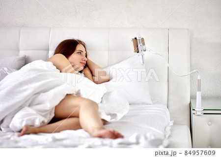 Smartphone holder with clamp is fixed on bedside table, woman is watching video of lie in bed. Smartphone holder with clamp is fixed on bedside table, woman is watching video of lie in bed. 84957609