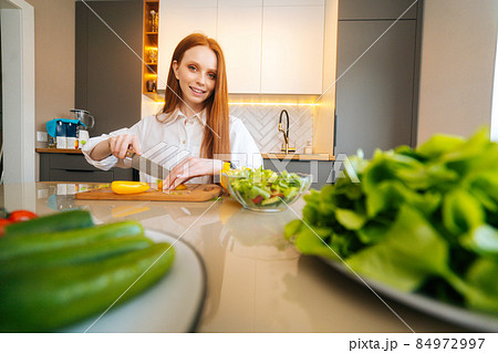 Portrait of young redhead woman cutting fresh yellow bell pepper preparing food salad sitting at table in modern kitchen room, looking at camera. Portrait of young redhead woman cutting fresh yellow bell pepper preparing food salad sitting at table in modern kitchen room, looking at camera. 84972997