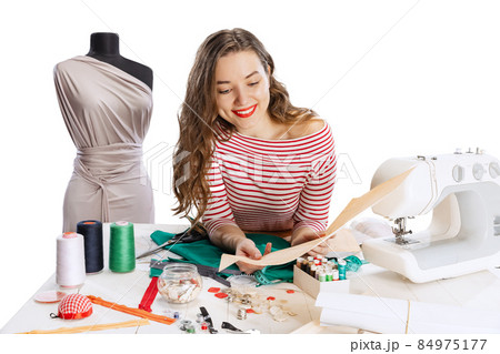 Cheerful beautiful young girl, seamstress, dressmaker working with fabric, scissors and sewing machine isolated on white background. Concept of job, hobby Cheerful beautiful young girl, seamstress, dressmaker working with fabric, scissors and sewing machine isolated on white background. Concept of job, hobby 84975177
