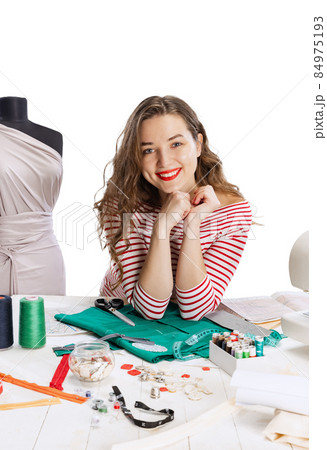 Smiling cute young girl, seamstress, dressmaker working with fabric, scissors and sewing machine isolated on white background. Concept of job, hobby Smiling cute young girl, seamstress, dressmaker working with fabric, scissors and sewing machine isolated on white background. Concept of job, hobby 84975193