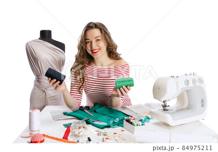 Cheerful beautiful young girl, seamstress, dressmaker working with fabric, scissors and sewing machine isolated on white background. Concept of job, hobby 84975211