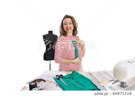 Cheerful beautiful young girl, seamstress, dressmaker working with fabric, scissors and sewing machine isolated on white background. Concept of job, hobby 84975228