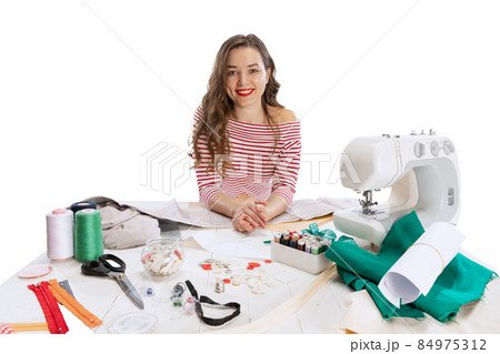 Cheerful beautiful young girl, seamstress, dressmaker working with fabric, scissors and sewing machine isolated on white background. Concept of job, hobby Cheerful beautiful young girl, seamstress, dressmaker working with fabric, scissors and sewing machine isolated on white background. Concept of job, hobby 84975312