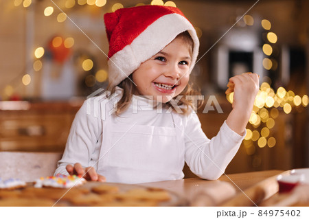 Adorable little girl in santa hat eating homemade gingerbread and laughing. Happy little kid preparing for Christmas. Background of fairy lights bokeh 84975402
