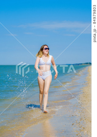 A young girl runs along the sandy sea beach along the surf. Splashes of sea water. The girl is wearing a white swimsuit and sunglasses. Freedom and carelessness. Outdoor fitness. Summer sunny day 84979803