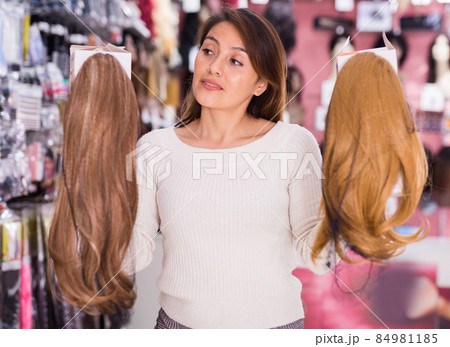 Smiling girl choosing wigs from natural hair in store Smiling girl choosing wigs from natural hair in store 84981185