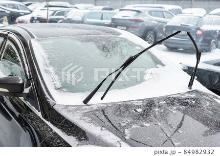 Close-up detail view of black modern car with raised up windshield wiper blades to prevent icing in cold frost winter day. Vehicle windscreen covered by whote snow parked on city street outdoors 84982932