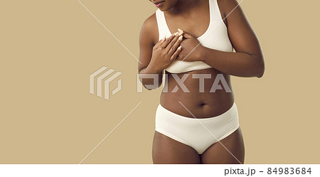 Beautiful plus size young black woman in underwear touches her chest when she feels strong pain standing isolated on beige color banner background. Breast cancer and heart attack awareness concept 84983684