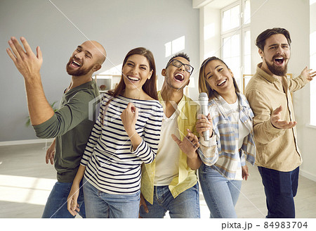 Young, cheerful and funny friends with microphone sing loudly and laugh while playing karaoke at home party. Close up of people in casual clothes. Concept of friendship, home entertainment and people. 84983704