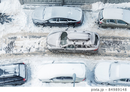 Top above aerial view of modern suv crossover car with roof rack box trunk driving through vehicles parked on city street covered by deep snow on clod frosty winter day. Apartment building driveway 84989148