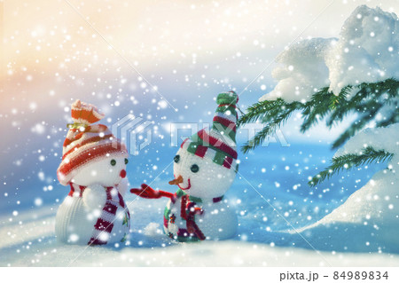 Two small funny toys baby snowman in knitted hats and scarves in deep snow outdoors near pine tree branch. Happy New Year and Merry Christmas greeting card. 84989834