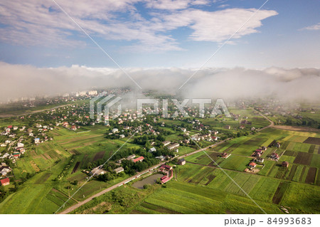 Aerial view of white clouds above a town or village with rows of buildings and curvy streets between green fields in summer. Countryside landscape from above. Aerial view of white clouds above a town or village with rows of buildings and curvy streets between green fields in summer. Countryside landscape from above. 84993683