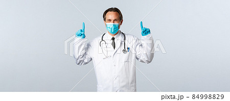Covid-19, preventing virus, healthcare workers and vaccination concept. Optimistic confident smiling doctor in white coat, medical mask and gloves pointing fingers up, advice or recommend product Covid-19, preventing virus, healthcare workers and vaccination concept. Optimistic confident smiling doctor in white coat, medical mask and gloves pointing fingers up, advice or recommend product 84998829