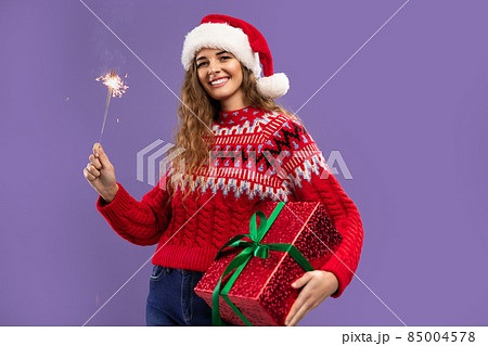 Happy beauty girl wears Santa hat holds gift box and sparkler on purple background. Christmas party Happy beauty girl wears Santa hat holds gift box and sparkler on purple background. Christmas party 85004578