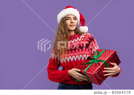 Beautiful smiling girl wears Santa hat holds red gift box on purple background. Christmas party Beautiful smiling girl wears Santa hat holds red gift box on purple background. Christmas party 85004579