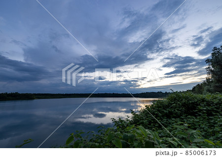 a beautiful blue lake with green tree in the foreground under a dramatic blue dusk sky 85006173