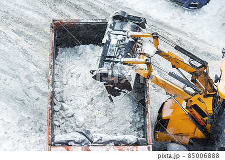Tractor loader machine uploading dirty snow into dump truck. Cleaning city street, removing snow and ice after heavy snowfalls and blizzard. Snowplow outdoors clean pavement sidewalk road driveway 85006888