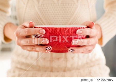 Woman's hands with french manicure and candy cane pattern on the nails. Woman with beautiful manicure holding big red knitted cup. The concept of cozy Christmas holidays and New Year. 85007905