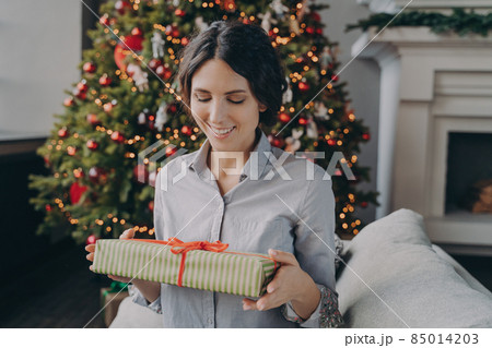 Smiling european woman sitting on sofa in room decorated for xmas holidays holding Christmas present 85014203