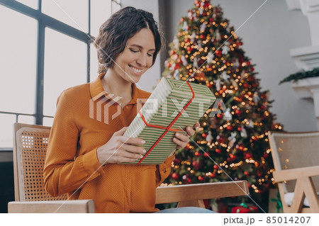 Beautiful happy Italian woman sitting with gift next to festively decorated Xmas tree at home 85014207