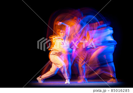 Excited young girl in white costume dancing hip-hop dance isolated on dark background at dance hall in neon mixed light. 85016285